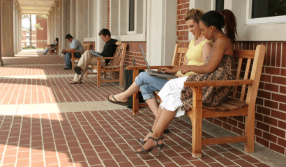 College students sit outside on benches along a wall while studying.
