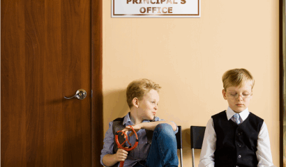 Two schoolboys sitting next to principal's office. They have different behavior and clothes.