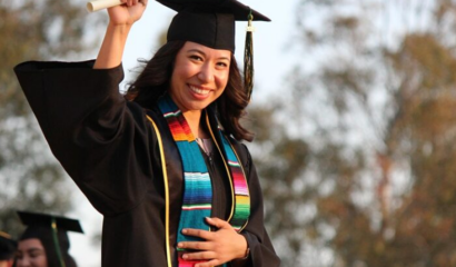 A Latina college graduate stands in her cap and gown, raising her diploma in the air.